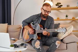 auf dem Sofa. Akustikgitarre spielen. Mann in Freizeitkleidung ist drinnen foto