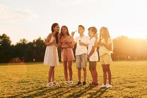 von Sonnenlicht beleuchtet. Stehen auf dem sportlichen Feld. Eine Gruppe glücklicher Kinder ist tagsüber im Freien foto