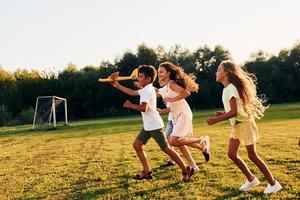 Laufen mit Spielzeugflugzeug. Eine Gruppe glücklicher Kinder ist tagsüber draußen auf dem Sportplatz foto