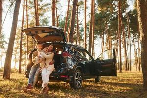 ans Auto gelehnt. glückliche familie von vater, mutter und kleiner tochter ist im wald foto