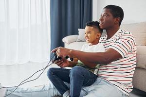 Verwenden von Joysticks zum Spielen von Videospielen. Afroamerikanischer Vater mit seinem kleinen Sohn zu Hause foto