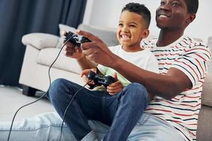 Videospiele spielen. Afroamerikanischer Vater mit seinem kleinen Sohn zu Hause foto