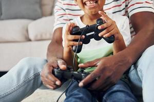 mit Joysticks in den Händen. Afroamerikanischer Vater mit seinem kleinen Sohn zu Hause foto