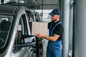 mit Kiste in der Hand. lieferbote in uniform ist drinnen mit auto und mit bestellung foto