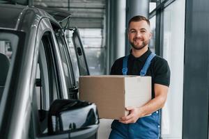 mit Kiste in der Hand. lieferbote in uniform ist drinnen mit auto und mit bestellung foto