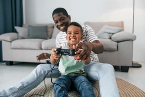Verwenden von Joysticks zum Spielen von Videospielen. Afroamerikanischer Vater mit seinem kleinen Sohn zu Hause foto