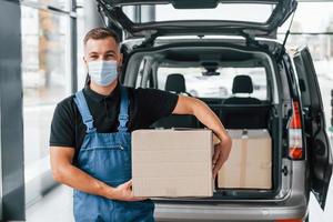 in Maske. lieferbote in uniform ist drinnen mit auto und mit bestellung foto