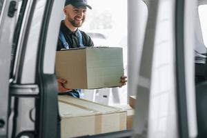 fröhlicher Arbeiter. lieferbote in uniform ist drinnen mit auto und mit bestellung foto