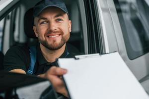 fröhlicher Arbeiter. lieferbote in uniform ist drinnen mit auto und mit bestellung foto