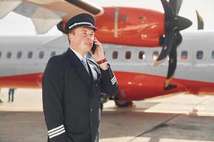 telefonieren. pilot in formeller schwarzer uniform steht draußen in der nähe des flugzeugs foto