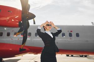weit weg schauen. junge stewardess in formeller schwarzer kleidung steht draußen in der nähe des flugzeugs foto