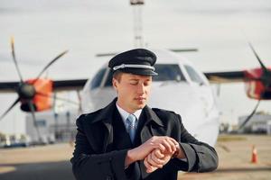 Überprüfung der Zeit. pilot in formeller schwarzer uniform steht draußen in der nähe des flugzeugs foto