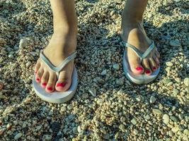 weiße Gummipantoffeln stehen auf dem Sand. Meeresschuhe. Schiefer für Spaziergänge entlang der Küste. weibliche füße mit rosa pediküre gehen in einem warmen land foto