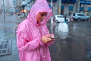 Eine junge Frau in einem Regenmantel schaut auf das Smartphone foto