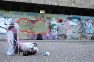 mehrere gebrauchte sprühdosen mit rosa und weißer farbe liegen auf dem asphalt vor dem stehenden kerl vor einer bemalten wand in farbigen graffiti-zeichnungen foto