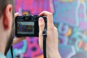 Ein junger Graffiti-Künstler fotografiert sein fertiges Bild an der Wand. Der Typ verwendet moderne Technologie, um eine farbenfrohe abstrakte Graffiti-Zeichnung aufzunehmen. Konzentrieren Sie sich auf das Aufnahmegerät foto