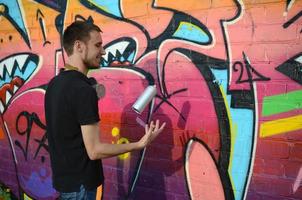 Der junge Graffiti-Künstler mit Gasmaske am Hals wirft seine Sprühdose gegen bunte rosafarbene Graffiti an der Ziegelwand. Street Art und zeitgenössischer Malprozess foto