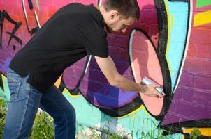 Der junge Graffiti-Künstler mit Rucksack und Gasmaske am Hals malt bunte Graffiti in rosa Tönen auf die Ziegelwand. Street Art und zeitgenössischer Malprozess foto