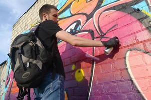 Der junge Graffiti-Künstler mit Rucksack und Gasmaske am Hals malt bunte Graffiti in rosa Tönen auf die Ziegelwand. Street Art und zeitgenössischer Malprozess foto