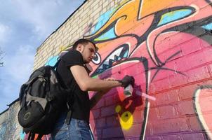 Der junge Graffiti-Künstler mit Rucksack und Gasmaske am Hals malt bunte Graffiti in rosa Tönen auf die Ziegelwand. Street Art und zeitgenössischer Malprozess foto