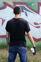 Ein junger Hooligan mit einer Spraydose steht an einer Betonwand mit Graffiti-Gemälden. illegales vandalismuskonzept. Straßenkunst foto