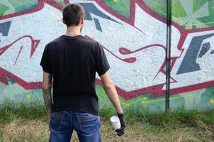 Ein junger Hooligan mit einer Spraydose steht an einer Betonwand mit Graffiti-Gemälden. illegales vandalismuskonzept. Straßenkunst foto