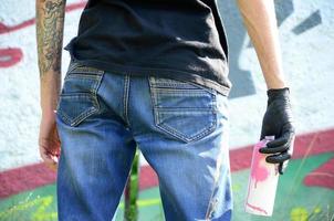 Ein junger Hooligan mit einer Spraydose steht an einer Betonwand mit Graffiti-Gemälden. illegales vandalismuskonzept. Straßenkunst foto