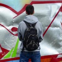 Ein junger Graffiti-Künstler mit einer schwarzen Tasche betrachtet die Wand mit seinem Graffiti an einer Wand. Street-Art-Konzept foto