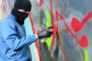 Ein junger Rowdy mit verstecktem Gesicht malt Graffiti auf eine Metallwand. illegales vandalismuskonzept foto