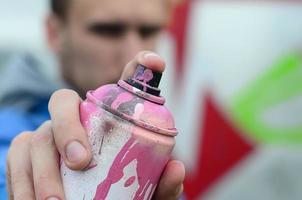 Ein junger Graffiti-Künstler in einer blauen Jacke hält vor dem Hintergrund einer farbigen Graffiti-Zeichnung eine Farbdose vor sich. Straßenkunst- und Vandalismuskonzept foto