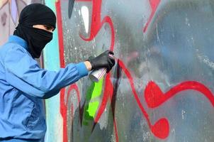 Ein junger Rowdy mit verstecktem Gesicht malt Graffiti auf eine Metallwand. illegales vandalismuskonzept foto