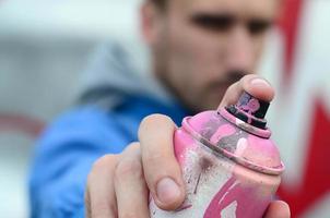 Ein junger Graffiti-Künstler in einer blauen Jacke hält vor dem Hintergrund einer farbigen Graffiti-Zeichnung eine Farbdose vor sich. Straßenkunst- und Vandalismuskonzept foto