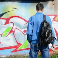 Ein junger Graffiti-Künstler mit einer schwarzen Tasche betrachtet die Wand mit seinem Graffiti an einer Wand. Street-Art-Konzept foto