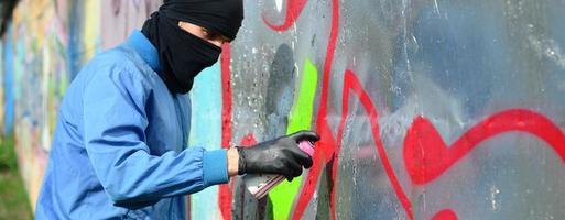 Ein junger Rowdy mit verstecktem Gesicht malt Graffiti auf eine Metallwand. illegales vandalismuskonzept foto