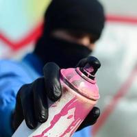 Ein junger Graffiti-Künstler in blauer Jacke und schwarzer Maske hält eine Farbdose vor sich vor dem Hintergrund einer farbigen Graffiti-Zeichnung. Straßenkunst- und Vandalismuskonzept foto