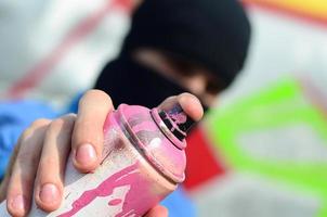 Ein junger Graffiti-Künstler in blauer Jacke und schwarzer Maske hält eine Farbdose vor sich vor dem Hintergrund einer farbigen Graffiti-Zeichnung. Straßenkunst- und Vandalismuskonzept foto