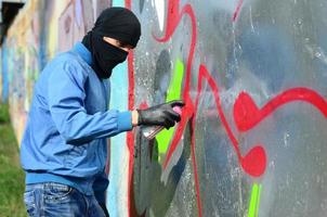 Ein junger Rowdy mit verstecktem Gesicht malt Graffiti auf eine Metallwand. illegales vandalismuskonzept foto