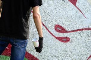 Ein junger Hooligan mit einer Spraydose steht an einer Betonwand mit Graffiti-Gemälden. illegales vandalismuskonzept. Straßenkunst foto