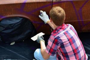 Ein junger rothaariger Graffiti-Künstler malt ein neues buntes Graffiti auf das Auto. Foto des Prozesses des Zeichnens eines Graffitis auf einem Auto, Nahaufnahme. das konzept der straßenkunst und des illegalen vandalismus