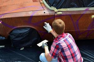 Ein junger rothaariger Graffiti-Künstler malt ein neues buntes Graffiti auf das Auto. Foto des Prozesses des Zeichnens eines Graffitis auf einem Auto, Nahaufnahme. das konzept der straßenkunst und des illegalen vandalismus