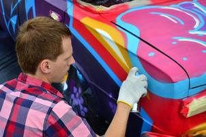 Ein junger rothaariger Graffiti-Künstler malt ein neues buntes Graffiti auf das Auto. Foto des Prozesses des Zeichnens eines Graffitis auf einem Auto, Nahaufnahme. das konzept der straßenkunst und des illegalen vandalismus