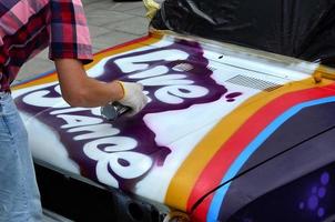 Ein junger rothaariger Graffiti-Künstler malt ein neues buntes Graffiti auf das Auto. Foto des Prozesses des Zeichnens eines Graffitis auf einem Auto, Nahaufnahme. das konzept der straßenkunst und des illegalen vandalismus