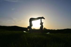 Silhouette zwei junge blonde Mädchen in Sportanzügen, die abends bei Sonnenuntergang auf einem malerischen grünen Hügel Yoga praktizieren. das konzept der übung und des gesunden lebensstils foto