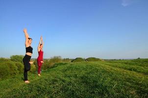 zwei junge blonde mädchen in sportanzügen praktizieren abends yoga auf einem malerischen grünen hügel unter freiem himmel. das konzept der sportausübung und der gesunden lebensweise foto