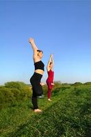 zwei junge blonde mädchen in sportanzügen praktizieren abends yoga auf einem malerischen grünen hügel unter freiem himmel. das konzept der sportausübung und der gesunden lebensweise foto