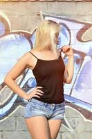 sexy kaukasisches blondes mädchen in jeansshorts und schwarzem tanktop, das tagsüber im freien gegen graffitiwand posiert foto