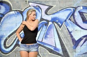 sexy kaukasisches blondes mädchen in jeansshorts und schwarzem tanktop, das tagsüber im freien gegen graffitiwand posiert foto