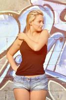 sexy kaukasisches blondes mädchen in jeansshorts und schwarzem tanktop, das tagsüber im freien gegen graffitiwand posiert foto