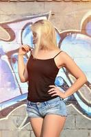 sexy kaukasisches blondes mädchen in jeansshorts und schwarzem tanktop, das tagsüber im freien gegen graffitiwand posiert foto