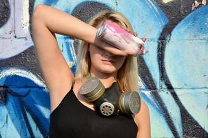 Junge und schöne sexy Mädchen-Graffiti-Künstlerin mit Gasmaske am Hals, die seine Augen mit einer Sprühdose versteckt, die auf einem Wandhintergrund mit einem Graffiti-Muster in blauen und violetten Tönen steht foto